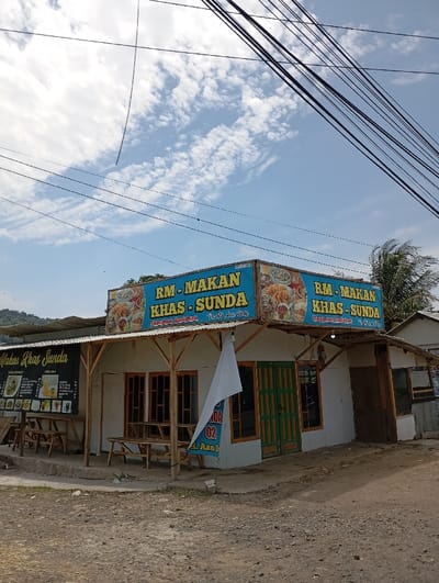 A small restaurant building, with a blue and white sign reading "RM-MAKAN KHAS-SUNDA" in Indonesian, located in Kecamatan Kutawaringin, Indonesia. The restaurant is made of wood and has a white awning covering the front. There are three wooden tables and chairs on the ground in front of the building. A white banner is hanging on the wall. The building is located on a dirt road. There are numerous power lines stretching overhead. There is a bright, clear sky with clouds.  The restaurant is situated on a small hill and there are trees in the background.  A second sign is visible on the right side of the building, also reading "RM-MAKAN KHAS-SUNDA". The restaurant appears to be closed.