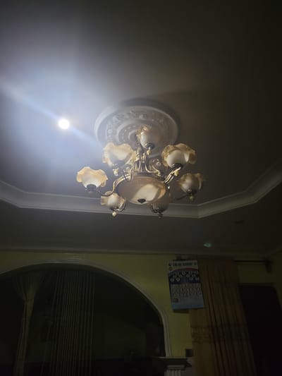 The image shows a dimly lit interior in Madalla, Nigeria. A decorative chandelier with multiple lights is affixed to a ceiling with decorative molding. A calendar is mounted on a wall. An arched doorway leads to another space with draped fabric or curtains.
