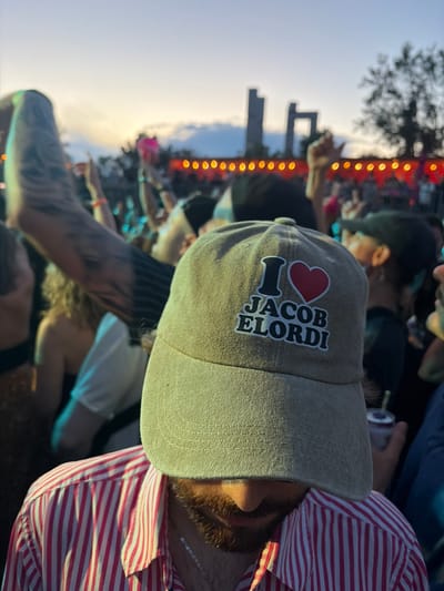 A person is seen wearing a tan baseball cap with a patch that says "I ❤️ JACOB ELORDI". They are also wearing a pink and white striped shirt. The person has a beard and is looking down. The background is blurred and appears to be a crowd at an outdoor event, possibly a concert, with lights and buildings visible in the distance. The sky is overcast with a sunset or sunrise glow. The location is not discernible from the image itself.