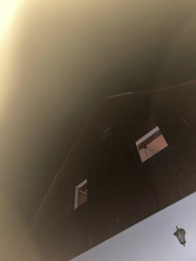 This is a low-angle, indoor shot of the attic space of a dark-colored building, possibly a house, located in Jablonné nad Orlicí, Czechia. The scene is dimly lit, with a soft, ambient light source coming from the upper left of the frame, creating a gradient of light and shadow across the image.

The primary subject is the angled roof structure of the attic, which is constructed from dark wood panels. Several architectural features are visible within this space. Two small, rectangular windows are cut into the roof. The left window is partially obscured by shadow but appears to be empty. The right window is more clearly visible, revealing an interior space with a reddish-pink wall and exposed wooden beams.

In the upper portion of the image, there are some exposed wooden beams and possibly white-painted structural elements. A thin, reflective line, possibly a wire or a piece of metal, is visible diagonally across the dark wooden surface of the roof.

In the lower right corner of the image, a vintage-style black lantern is hanging. It has a dark, ornate metal frame and clear glass panels. The lantern appears to be decorative and is the only clear light fixture visible in the frame.

The overall impression is one of an unfinished or seldom-used attic space, with a focus on the structural elements and a hint of interior detail through the windows. The lighting and angle suggest the photo was taken by someone looking upwards into the attic.