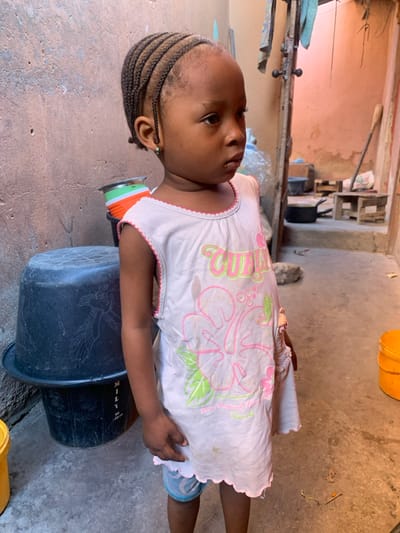 A young Black child, approximately 2-4 years old, stands facing slightly right of the camera, looking straight ahead with a neutral expression. The child has cornrow braids pulled back from the forehead and wears a light-colored, sleeveless dress with pink scalloped trim and a graphic featuring a pink hibiscus flower and green leaves. Light-blue patterned shorts are visible beneath the dress. A small green earring is present in the left earlobe.
The scene is an outdoor or semi-enclosed urban space, likely a courtyard or alleyway in Okene, Nigeria. The immediate background features a rough, textured concrete or stucco wall, painted an aged beige/light brown, exhibiting patches of wear and discoloration. To the child's left, two dark plastic buckets are stacked, the upper one inverted. Behind the child's right shoulder, a partial view of an open doorway or archway reveals a brighter, reddish-orange painted wall beyond, suggesting an interior or connecting space. Various utilitarian objects are visible in the background through this opening, including what appears to be a black cooking pot, small wooden stools or pallets, and hanging fabric. The ground is a flat, grey concrete surface with minor debris. To the far right, a stack of two yellow plastic buckets is partially visible.