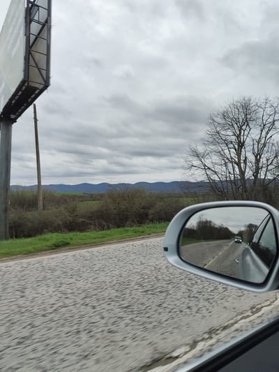 The image depicts a view from a moving vehicle, likely a car, on a road near Mramoren, Bulgaria. In the foreground is a textured asphalt road. To the left, a roadside with green grass and scrub vegetation is visible, along with a tall billboard structure on a metal pole. In the middle ground, a line of scrubby bushes and trees leads to a range of rolling hills on the horizon. The sky is overcast with gray clouds. A side view mirror of the vehicle reflects the road behind, showing another car in the distance. A bare tree stands to the right of the mirror.
