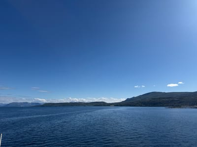 This media file is a landscape photograph taken from a boat on a body of water, likely a fjord, in Hierenjárgga - Korsnes, Norway. The scene is dominated by the deep blue expanse of the water, which has a slightly rippled surface, reflecting the clear blue sky above. In the distance, a lush, green, and hilly coastline stretches across the frame. The land is densely covered with trees and shrubs, with some rocky outcrops visible along the water's edge. The terrain rises into rolling hills and mountains, some of which are partially obscured by clouds. The sky is a vivid, cloudless blue for the most part, with a few wispy white clouds scattered near the horizon. The bright sunlight suggests it is daytime, likely mid-morning or afternoon, and the weather appears to be clear and fair. There is a sense of tranquility and natural beauty conveyed by the serene water and the majestic, tree-covered landscape. The image is composed to emphasize the vastness of the water and sky, with the coastline serving as a scenic backdrop. The overall impression is one of a peaceful day in a scenic, natural environment.