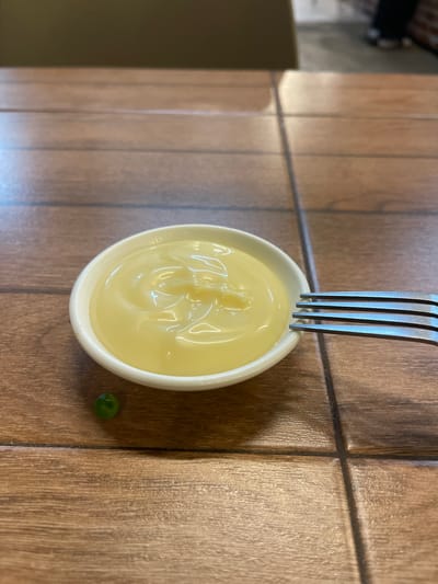 A close-up, eye-level shot shows a small white ramekin filled with creamy yellow mayonnaise, positioned on a wooden table. A single green pea rests to the left of the ramekin. A metal fork, with its tines partially submerged in the mayonnaise, is visible to the right of the ramekin. The table surface is composed of wooden planks, with visible grout lines between them. The background is blurred, suggesting an indoor environment, possibly a restaurant or home.