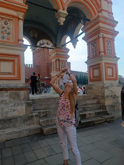 This detailed image captures an outdoor scene in Elektrougli, Russia, featuring a fair-skinned woman in the foreground engaged in photography. She has long blonde hair, styled back, and is wearing a white sleeveless top with a colorful floral pattern of red, pink, blue, and green flowers, paired with white ripped jeans. Bracelets are visible on her left wrist, and a necklace with a small pendant adorns her neck. She holds a dark-colored smartphone with both hands, tilting her head sharply upwards and backward, aiming the device towards the ornate archway directly above her. A dark-colored backpack with a striped pattern is worn on her back. She stands on a paved surface of dark grey square tiles, at the base of worn, light grey stone steps.

The woman is positioned within a large, decorative architectural structure composed of robust, light-colored stone pillars and multiple archways, accented with prominent orange-red borders and details. The inner ceilings of the larger archways are painted light blue with scattered gold and blue floral or star-like patterns. Underneath a smaller arch, a darker, intricate pattern with a central circular motif containing a cross-like symbol is visible. Vertical panels on the pillars display vibrant floral designs in red, blue, and green within swirling vine motifs, enclosed in square frames. Other panels feature plain orange-bordered squares. Dark, horizontal metal support bars extend between the pillars.

In the mid-ground and background, visible through the archways, are significant red brick walls of a fortress-like structure, featuring battlements along their top edge. A distinct red brick tower with a green-domed roof is partially visible through one archway. Several dark green coniferous trees stand behind the red walls. Numerous other people are scattered throughout the background, walking and standing, indicating a public and active environment. The sky above is clear and light blue, suggesting bright, fair weather conditions.
