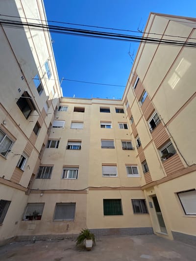 This media file captures an upward-looking view of a multi-story building, likely an apartment complex, on a clear and sunny day in Castelló De La Plana, Spain. The building's facade is painted a pale yellow, with numerous windows of varying sizes and styles. Some windows have protective bars, while others feature blinds or shutters.
The architecture appears functional, with balconies and external overhangs creating a staggered effect on some levels. Electrical wires hang prominently in the foreground, suggesting an urban or semi-urban setting. The clear blue sky dominates a significant portion of the frame, indicating fair weather.
The composition draws the eye upwards, emphasizing the height of the building and the sheer number of windows. The lighting suggests it is daytime, possibly late morning or early afternoon, given the strong shadows cast by the overhangs. A lone potted plant on a small patio visible on the right side provides a touch of greenery against the muted tones of the building and pavement. No people are visible, and there is no discernible text in the image. The overall impression is one of everyday urban life in a Mediterranean climate.