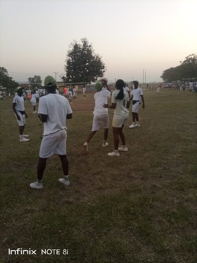 A group of people dressed in white shirts and shorts play a game of volleyball on a grassy field. The players are wearing green hats. A tree stands in the background, and another group of people sit watching the game. The sky is dusky. The image has the watermark "Infinix NOTE 8i" in the lower left corner.