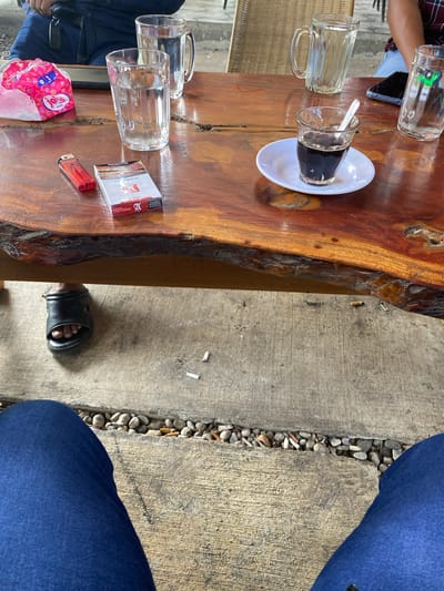 The image depicts a close-up view of a wooden table with several items on it, set in what appears to be an outdoor seating area with concrete flooring. A person's legs, clad in blue jeans, are visible at the bottom of the frame, suggesting they are seated at the table. A black sandal is worn on one foot. The table holds two glasses of water, a cigarette pack, a red lighter, and a small glass filled with a dark liquid, likely coffee, accompanied by a spoon on a saucer. Two additional glasses of water are partially visible in the background. The concrete floor shows scattered cigarette butts. The scene is likely located in Aluepiet, Indonesia, given the context provided.