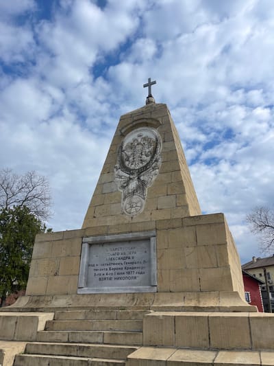 The image shows the Monument to the Liberators, specifically dedicated to the Battle of Nikopol, which took place in 1877 during the Russo-Turkish War. The monument is made of stone, featuring a cross at the top. A circular relief is placed on the monument, with a plaque below it bearing Cyrillic script. The inscription indicates that the monument commemorates the reign of Emperor Alexander II and the leadership of General Lieutenant Baron Krüdener during the capture of Nikopol on July 3rd and 4th, 1877. The sky is partially cloudy. Trees and a building are visible in the background.