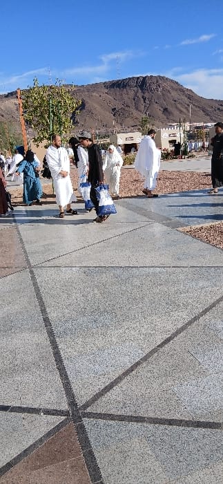The image displays an outdoor scene at the base of Mount Uhud in Medina, Saudi Arabia, under a clear blue sky. The foreground is dominated by a light grey, speckled concrete or tiled plaza featuring darker grey geometric dividing lines. Reddish-brown gravel areas flank the paved surface on the right and left. In the mid-ground, a group of approximately fifteen individuals is observed walking and standing on the plaza. Several men are dressed in white ihram garments, with visible sandals on their feet. A central male figure wears a black thobe and a dark cap, carrying two blue and white patterned tote bags in his left hand. Women are also present, one distinctly in a teal-colored abaya, others in white or partially obscured brown attire. To the left, a slender tree with sparse green foliage stands near a brown pole. In the right mid-ground, a series of low-rise buildings with light-colored, traditional architectural features and dark arched openings are visible, suggesting visitor amenities or shops. Tall, slender poles extend upwards from behind these structures. The background is dominated by the large, rugged, dark brown rocky mass of Mount Uhud, rising prominently against a clear, vibrant blue sky.