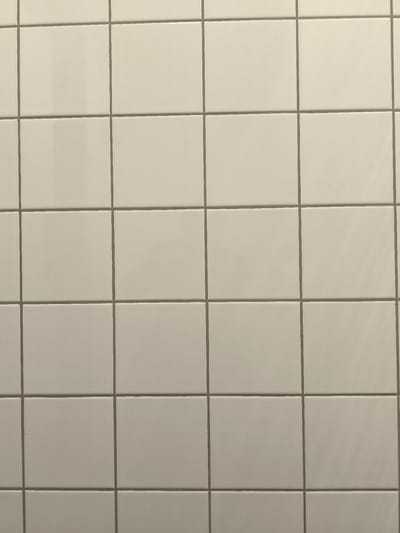 The image shows a close-up, full-frame view of a tiled wall. The tiles are square and a light, neutral color, likely beige or off-white, with visible grout lines that are a darker gray. The lighting appears to be even, with no strong shadows or highlights, suggesting an indoor environment. There are no people or distinct objects visible in the frame, only the repeating pattern of the tiles. The location in Komorniki, Poland is not discernible from the image itself.