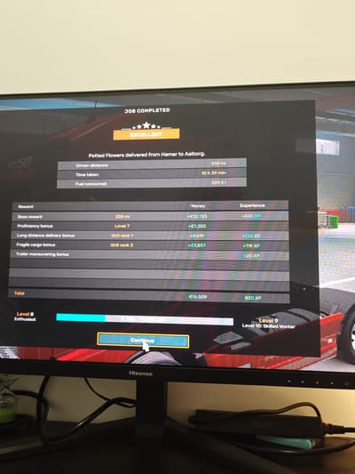 The image displays a "Job Completed" screen from a video game, likely a driving simulator like Euro Truck Simulator 2 or American Truck Simulator. The delivery was "Potted Flowers delivered from Hamar to Aalborg", a distance of 338 miles, taking 10 hours and 39 minutes, with a fuel consumption of 269.51. The job earned an "Excellent" rating.
The breakdown of the earnings includes:
* Base reward: +€12,733 (+545 XP)
* Proficiency bonus (Level 7): +€1,300
* Long distance delivery bonus (Skill rank 1): +€619 (+136 XP)
* Fragile cargo bonus (Skill rank 3): +€1,857 (+119 XP)
* Trailer maneuvering bonus: +€20 XP
The total earnings for the job were €16,509, along with 820 XP. The player is currently at Level 8 "Enthusiast" and has accumulated 7,788 of the 9,000 XP needed to reach Level 9 "Skilled Worker". A "Continue" button is displayed, allowing the player to move forward.
The image is captured from a monitor, identified as a Hisense brand. An ASUS device is situated below the monitor. An hourglass is present on the desk, partially visible.