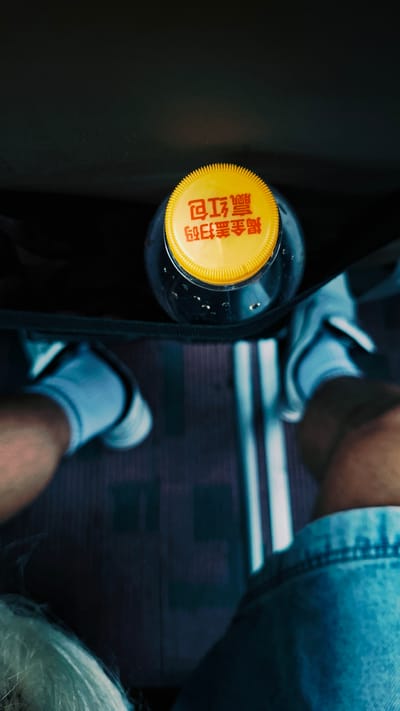 A top-down, high-angle shot shows a person sitting on a bus or train in Dongcheng District, China, with a bottle of tea and a yellow cap at their side. The subject wears light blue denim shorts, white sneakers, and grey socks. A black bag strap is across the bottle. The bottle is mostly out of focus. The background is dark with blue hues. The floor has a ridged pattern. The subject's hand can be seen in the bottom right of the image. The bottle cap has Chinese writing on it.