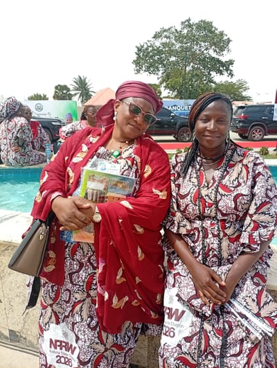 Two women are seated outdoors under bright daylight, likely at an event in Unguwan Sanusi, Nigeria. Both are smiling and looking at the camera.

The woman on the left wears a flowing red top with patterned sleeves and a matching headscarf, along with a patterned skirt. She has sunglasses on her head and a watch on her left wrist. She holds a light brown handbag and several brochures or pamphlets, one displaying text "MAUWE MOHAMMED" and images of buildings. A white patch on her skirt reads "NPRW Kaduna State Chapter 2026."

The woman on the right wears a V-neck dress made of the same patterned fabric as the first woman's skirt, featuring red, white, and brown tones. Her hair is styled in dark braids with hints of reddish-brown. She holds a folded item, possibly a piece of fabric or paper, which has text "VERITABLE BLOCK PRINTS NEW CHIG" and "RW Date Chapter 26" visible on a white patch.

In the background, a turquoise-colored fountain or small pool is visible. Other attendees, also dressed in patterned attire, are seated around the fountain. Further back, black SUVs are parked, and a white banner is partially visible with text such as "sinning Of" and "BANQUET." Large trees and a clear sky indicate an outdoor gathering or banquet setting.