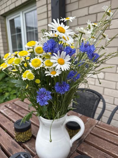 The image depicts a close-up view of a bouquet of wildflowers, including daisies and cornflowers, arranged in a white ceramic pitcher. The pitcher sits on a rustic wooden table outdoors. In the background, a brick wall and a window of a building are visible, along with the silhouette of outdoor furniture. There are no people visible in the image. The scene is set in a garden or patio area.