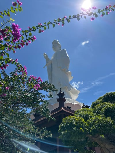 This is a low-angle, vertical shot of a large white statue against a bright blue sky with scattered white clouds. The statue appears to be a person, possibly female, with flowing robes and outstretched arm. The perspective makes the statue seem immense, towering over the viewer. In the foreground and framing the shot on the left and right are vibrant green trees and branches laden with bright purple flowers. The sun is visible on the left side of the image, casting a lens flare and bright light. The scene appears to be outdoors on a sunny day. The foliage and flowers suggest a warm climate and possibly a tropical or subtropical setting. The architectural elements partially visible behind the statue, with decorative wooden trim, hint at a temple or significant cultural structure. The overall mood is serene and awe-inspiring, with the natural elements adding a touch of beauty and vibrancy to the majestic statue. The location context provided is Thôn 5, Vietnam.