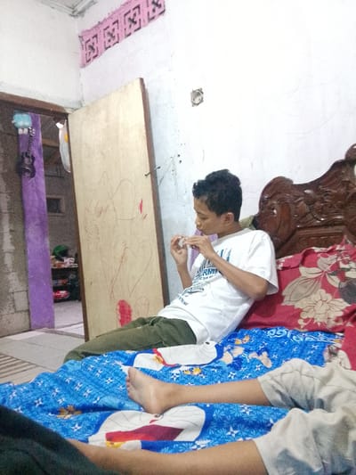 In an indoor residential setting, possibly a bedroom, in Cisarua, Indonesia, a young person with dark, curly hair is seated on a bed. They are wearing a white t-shirt and green pants. The individual is holding what appears to be a cigarette or a similar rolled item to their lips, with their hands engaged in the action. The bed is covered with a blue patterned blanket, and a portion of another person's leg, wearing light-colored shorts, is visible at the bottom of the frame, indicating the presence of at least two people.
The environment includes a wooden headboard of a bed, a partially open door with drawings on it, and a wall with some decorative elements at the top. To the left, a purple-colored column or wall section is visible, with a stringed instrument (possibly a guitar) hanging on it. Shelving units with various items can be seen through the open doorway. The lighting suggests an indoor space with ambient light. The overall impression is one of a casual, domestic scene.