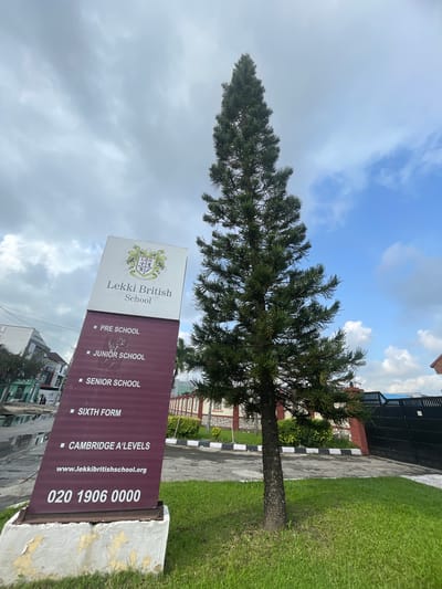 The image shows a sign for Lekki British School in Moba, Nigeria, with information about the school's various levels and contact details. A tall evergreen tree stands nearby. Beyond the sign, there is a glimpse of the urban landscape, which includes buildings and other signs of city life. The sky is partly cloudy. The foreground shows a lawn and a paved area.