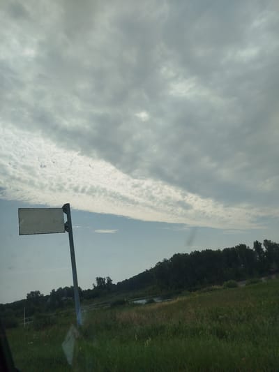 An overcast sky fills the upper portion of the image, with layered clouds creating a sense of depth. Below the sky, a dark line of trees forms a distant horizon. In the foreground, a grassy field stretches out, with a road visible in the mid-ground, bordered by a body of water. A metal signpost stands in the left foreground, with a blank white rectangular sign attached to it. The signpost is angled slightly to the right. The overall impression is of a rural landscape under a cloudy sky.