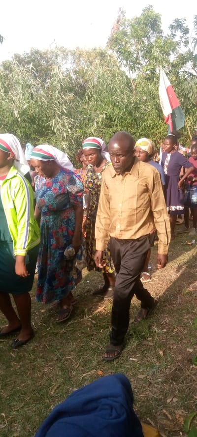 The image shows a group of people standing in a grassy area in Burundu, Kenya. The coordinates provided (latitude: 0.464, longitude: 34.74) place the location in the town of Burundu in the Bungoma County. There are seven people in the scene. The person in the foreground is a man wearing a light brown shirt, black pants, and black sandals. He is standing with his arms at his sides and looking toward the right side of the frame. To the man's left is a woman wearing a blue, red, and black patterned dress. She is holding a blue and white cloth in her hands. Behind her is a woman wearing a green and white headscarf and a black and yellow patterned dress. Behind the man is a woman wearing a blue and white headscarf and a black and white patterned dress. The woman on the far right is wearing a blue and white dress and a white headscarf. In the background, a flag can be seen hanging from a tree branch. The environment is green and grassy, with trees visible in the background. The image appears to be taken during the day. The lighting is bright and the colors are saturated. There are no indications of what event is occurring or why the group of people are gathered.