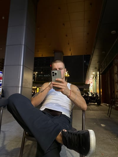 A man is seated on a chair, outdoors at night, in what appears to be a commercial district in Huangpu District, China. He is wearing a white tank top and dark pants, with black sneakers. The man is holding a smartphone and appears to be looking at it. The background shows a building with illuminated signage and several motorcycles parked outside. The lighting suggests it is nighttime, with artificial light sources illuminating the scene. The man has tattoos on his arms and is wearing bracelets.