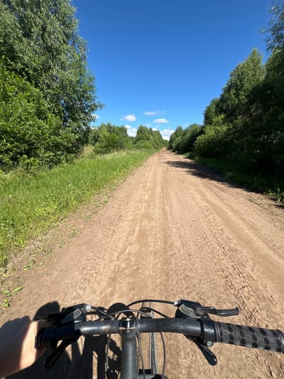 A first-person perspective shot captures a bicycle rider's view of a dirt road surrounded by lush green trees under a bright blue sky with scattered white clouds. The road ahead is well-traveled, showing tire tracks. The handlebars of the bicycle are visible in the foreground, with the rider's left hand holding the grip. The location is specified as Ural'skoye, Russia.