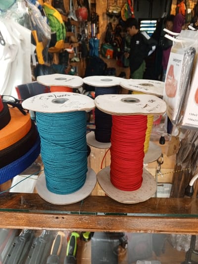 This image depicts the interior of a retail establishment, identified as being in Baza, Myanmar (Burma), viewed from slightly above a display counter. In the immediate foreground, a wooden frame supports a clear glass surface. Upon this surface, multiple large spools of cordage are arranged. Prominently featured are a teal spool on the left and a red spool to its right, both densely wound with rope. Other spools in the background contain black, yellow, and orange cordage. Each spool consists of a central cardboard core and circular cardboard ends, with handwritten markings visible on some top ends. To the far left, stacks of multicolored webbing or straps are visible, including orange, black, and blue sections, with a pair of black-framed eyeglasses resting on the orange stack.
The background reveals a densely packed store environment, displaying various merchandise typical of an outdoor equipment or general goods store. Items hanging on walls and racks include backpacks, clothing, and assorted accessories. Two individuals are present in the blurred mid-to-background. One, positioned centrally, is a male youth wearing a dark athletic jacket with white stripes (possibly an Adidas garment) and lighter pants, appearing to examine items on a display. Another person, partially obscured to the right, wears a dark top and is also in proximity to merchandise displays. The scene is illuminated by ambient light, highlighting the vibrant colors of the cordage and various products.