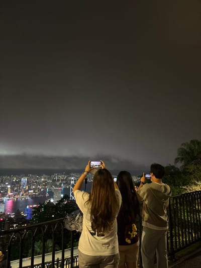 Three individuals are positioned along a railing, each holding a smartphone, apparently taking photographs of the city below. The first individual is a male wearing a gray tracksuit. The next two are women with long hair. One is wearing a tan-colored top and shorts with two yellow plush toys clipped to her shorts. The other is wearing a white shirt and gray shorts. The city is illuminated at night, possibly Hong Kong due to the distinct city lights and harbor scene. The fog or low clouds obscure parts of the city below. A portion of a black railing is visible along with trees and greenery.