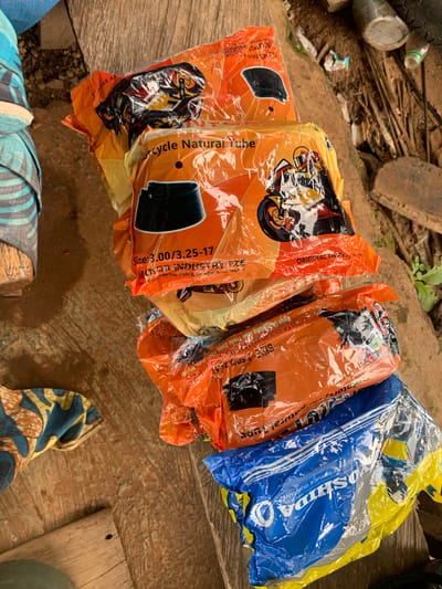 The image displays a close-up, high-angle view of several packaged items, likely motorcycle inner tubes, stacked on a rough wooden surface. The majority of the packages are made of clear orange plastic, revealing the contents. Each orange package features an illustration of a motocross rider on a motorcycle. Visible text on these orange packages includes "Motorcycle Natural Tube", "SIZE: 3.00/3.25-17", and "SIZE: 2.50/2.75-17". Further text mentions "MOTOR INDUSTRY FZE", "ORIGINAL QUALITY", and details like "Ogun Guangzhou Free Trade Zone, Ogun State, Nigeria", providing a specific location cue.
To the right, one package is distinctive in blue and yellow plastic, with the brand name "YOSHIDA" clearly visible, along with phrases like "PREMIUM QUALITY SERVICE GUARANTEE" and "MADE IN NIC".
The setting appears to be outdoors during daytime, indicated by the bright, natural lighting. The wooden surface beneath the packages is rough, and the background shows a cluttered, earthy area with debris, including a dark old tire, plastic bottles, and small pieces of wood, suggesting an informal outdoor environment. Parts of patterned fabric items, possibly clothing or bags, are visible in the bottom corners of the frame. The presence of the packages, particularly with the "Ogun State, Nigeria" text, implies a commercial or storage context, possibly in a market or roadside stall in Gbeleko, Nigeria. No people are visible in the frame.