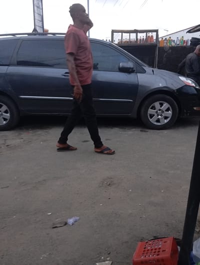 A man in a salmon-colored t-shirt and black pants walks past a dark gray minivan. He is seen in profile, with his right hand to his ear, as if on the phone. The man is wearing brown and black flip-flops. The minivan has a roof rack and is parked on a dirt or gravel road. In the background, there is a structure with shelves holding various bottles, possibly for sale. Another person, wearing dark clothing, is partially visible to the right of the minivan. The scene appears to be an outdoor market or roadside stall in Port Harcourt, Nigeria. A red plastic crate is visible in the lower right foreground. The sky is overcast.