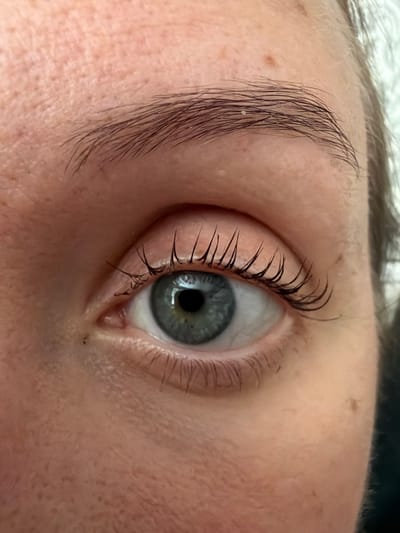 A close-up, high-angle, eye-level shot captures a detailed view of a human eye. The eye is open, revealing a blue iris with a dark pupil at the center. Long, dark eyelashes frame the eye. The skin surrounding the eye is fair with visible pores and a few small moles. The eyebrow above the eye is dark and appears naturally shaped. The lighting is bright, highlighting the textures of the skin and the details of the eye. The background is slightly blurred, suggesting a shallow depth of field. There are no discernible objects or structures indicating a specific location within Bila Tserkva, Ukraine.