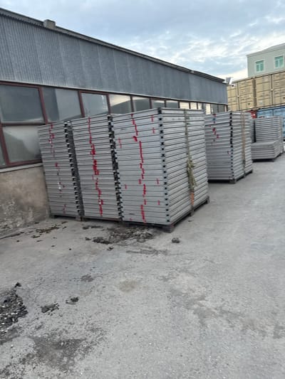In Baku City, Azerbaijan, a collection of stacked metal panels sits outdoors on a concrete surface. The panels are uniformly gray and bound together by straps, with red markings visible on some of the stacks. In the background, a corrugated metal building with large windows stands to the left, while shipping containers, some yellow and others blue, are visible to the right. The sky above is overcast. Debris, including small rocks and what appears to be dried mud, is scattered on the concrete ground in the foreground. No people are present in the scene.