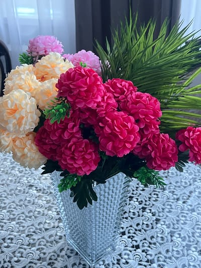 In a bright interior, a clear glass vase filled with a vibrant artificial flower arrangement sits on a white lace tablecloth. The bouquet features a mix of fuchsia, peach, and light pink flowers with prominent, layered petals. Lush green artificial foliage is interspersed throughout the arrangement, with longer, palm-like fronds rising from the back. The background shows a blurred window with sheer curtains on the left and a dark curtain on the right. The scene evokes a decorative indoor setting, potentially a home or a reception area, with no specific identifiable landmarks linking it to Kaspiysk, Russia.