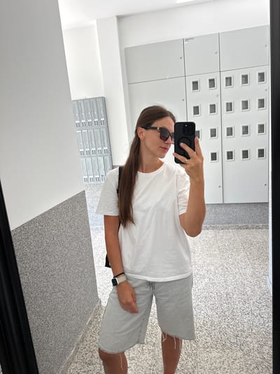 A woman is taking a mirror selfie in a well-lit, modern hallway. She is wearing a white t-shirt, light-wash denim shorts, sunglasses, and a watch on her left wrist. She is holding a black smartphone in her right hand, which is being used to take the picture. Behind her, the hallway features a speckled gray floor and a wall covered with lockers or storage units. To her left, there appears to be a textured gray wall with a diagonal white partition. The lockers in the background are arranged in a grid pattern and have small, square openings. The overall environment suggests an interior space, possibly a building with communal facilities.