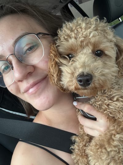A close-up selfie shows a woman with glasses and a brown poodle. The woman is holding the dog, which is resting its head on her shoulder. They appear to be in a car, with a seatbelt visible across the woman's chest. The background is blurred, but suggests an outdoor setting. The image was taken in Pervomaysky, Russia.