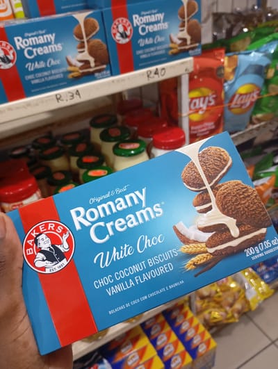 A person is holding a blue box of "Romany Creams White Choc" biscuits in a shop. Behind the box are shelves filled with various products, including jars with red and green lids, Lay's chips, and other boxes of the same biscuits priced at R.34 and R.40. The biscuits are chocolate coconut flavor with vanilla. The location is likely a shop in Johannesburg, South Africa, given the product and pricing.