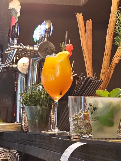 The image shows a bar scene with several drinks and bar tools. In the foreground, there is a glass with an orange-colored drink garnished with a slice of orange and a cherry. Next to it, there is a clear glass with ice and a green drink, possibly a mojito, with mint leaves. Behind these drinks, a collection of black straws is neatly arranged in a holder.

To the left, a beer tap system is visible, with chrome handles and a decorative figurine on top of one of the taps. A small potted rosemary plant adds a touch of greenery to the scene. In the background, several cinnamon sticks stand upright, adding a rustic element. The overall environment appears to be a bar with a dark background, possibly indicating an indoor setting.

The presence of a beer tap system and the general bar setup suggest this location is likely a bar or restaurant. Without more specific visual cues such as signage or distinctive architectural features, it is impossible to definitively pinpoint the city of Iquique, Chile. However, if we assume the location is Iquique, this scene depicts a typical bar setting within that city.