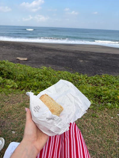 In the foreground, a person's left hand, adorned with a light pink nail polish, holds a pastry wrapped in white paper. The pastry is a rectangular shape, covered in sesame seeds. The paper wrapping displays text that reads "Your everyday Nordic toast" and "copenhagen". The person is wearing a red and white striped skirt. In the background, a dark sand beach meets the ocean, with gentle waves rolling towards the shore under a partly cloudy sky. Lush green vegetation with small yellow flowers grows between the beach and the grassy area where the person is situated. The overall scene captures a moment of relaxation by the sea.