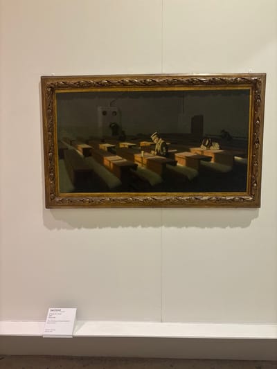 The media file presents a photograph of a framed painting displayed on a light-colored wall in what appears to be an art exhibition space, likely a museum or gallery.
**Subjects:** The central subject is a painting titled "The Christmas of those left behind." It depicts a somber interior scene with several elderly men seated in rows on benches or pews. Their figures are mostly shown from the back or in profile, appearing contemplative or resigned. The men are dressed in dark clothing, blending with the overall dark palette of the painting.
**Objects:** Within the painting, the main objects are the benches and the figures of the men. A dramatic light source, unseen but implied, casts strong light and shadow patterns across the interior, highlighting elements like the tops of the benches and the men's heads, creating a striking chiaroscuro effect. The painting itself is housed in an ornate, golden, carved wooden frame. On the wall beside the painting, a white informational plaque is visible.
**Scene and Setting:** The painting's setting is a large, dimly lit indoor space, possibly a communal hall, dormitory, or refectory. The exhibition setting is a modern, brightly lit room with plain white walls.
**Activity or Event:** Within the painting, the men appear to be gathered, sitting quietly, perhaps in reflection or during a meal. The title, "The Christmas of those left behind," suggests an event related to the Christmas holiday, implying themes of solitude, hardship, or introspection among those who might be isolated or forgotten during such a time.
**Notable Details:** The painting's atmosphere is melancholic and introspective, created through its subdued colors and dramatic lighting. The strong contrast between light and deep shadow is a prominent artistic detail. The overall photograph is taken slightly off-angle, showing the painting and a portion of the surrounding wall. The location context of Venice, Italy, aligns with the "Biennale, 1903" text on the plaque, suggesting the painting was exhibited or acquired at the Venice Biennale.
**Visible Text:** The white informational plaque provides details about the artwork:
- **Artist:** Angelo Morbelli (Alessandria 1853 / Milano 1909)
- **Italian Title:** Il Natale dei rimasti
- **Year of Creation:** 1903
- **Medium (Italian):** Olio su tela
- **English Title:** The Christmas of those left behind
- **Medium (English):** Oil on canvas
- **Acquisition Information:** Acquisto/Purchase, Biennale, 1903