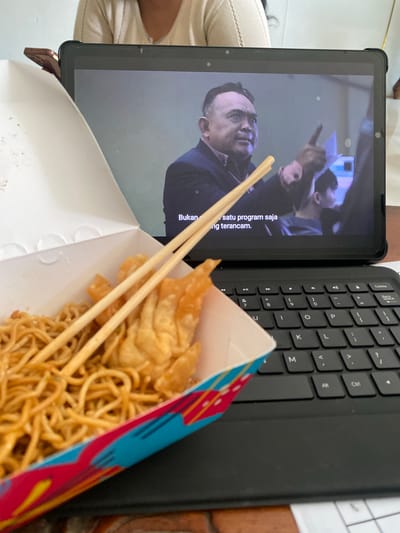 The image shows a close-up of a takeout box of noodles and fried wontons, with chopsticks resting on top, placed on a black keyboard. In the background, a tablet displays a video of a man speaking, pointing his finger. The scene appears to be indoors, likely in a home or office setting. The keyboard is a standard QWERTY layout, and the tablet is positioned behind the keyboard. The noodles and wontons are in a white paper box with a colorful, abstract design. The man on the tablet is wearing a dark jacket over a collared shirt. There are no discernible details in the image that would indicate a specific location in East Jakarta, Indonesia.