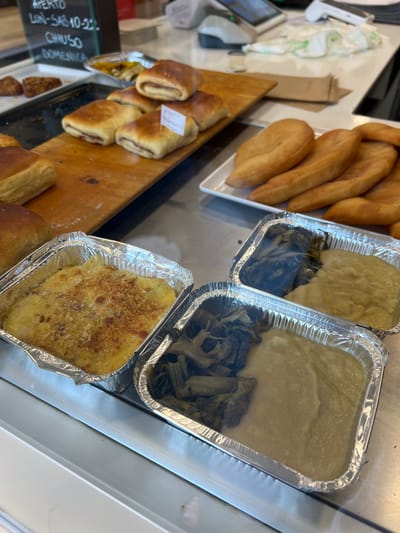 The image displays a close-up, eye-level view of food items arranged in a refrigerated display case, likely in a bakery or deli in Bari, Italy. The background shows a blurred retail environment with a cash register and a sign written in Italian that appears to indicate opening hours: "APERTO LUN-SAB 10-22: CHIMSO DOMENICA".
In the foreground, to the left, is a rectangular aluminum foil container filled with a golden-brown baked dish topped with breadcrumbs. Next to it, and slightly behind, is another similar container with two distinct sections: one filled with dark green cooked leafy vegetables, and the other with a pale yellow, mashed or pureed substance.
Further back and to the right, on a wooden board, are several rectangular pastries, some with visible reddish filling. These pastries are golden brown and appear to be freshly baked. To their right, on a white rectangular plate, are several pieces of fried dough, also golden brown and somewhat puffy.
The overall impression is one of ready-to-eat food items presented for sale. The lighting is bright, typical of a commercial establishment. There are no people clearly visible in the image.