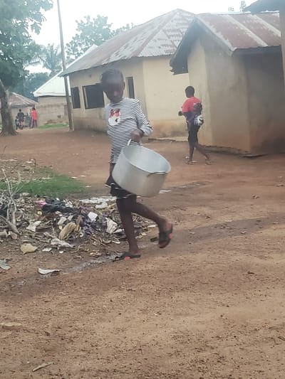 The image depicts a scene in Gumel, Nigeria, featuring a young girl walking across a dirt path while carrying a large metal pot. The girl, wearing a horizontally striped long-sleeved shirt and a dark skirt, is in motion, her body angled towards the right of the frame. She is barefoot, wearing flip-flops on her feet. To her left, a pile of assorted trash, including plastic bags and debris, lies on the ground.
Behind the girl, another individual, a person wearing a red t-shirt and dark pants, is visible in the mid-ground, appearing to walk away from the camera. They are carrying something on their back. In the background, several buildings with corrugated metal roofs are present, suggesting a residential area. The overall environment appears somewhat unkempt, with the dirt path and scattered refuse. Trees and foliage are visible in the upper left portion of the image. The sky is overcast, contributing to a diffused light.