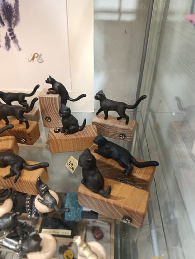 The image displays a collection of miniature black cat figurines arranged on wooden blocks within a glass display case, likely in a shop. The figurines vary in pose, with some standing, some walking, and one sitting. Some of the wooden blocks have small, decorative paw prints carved into them. A price tag with "39,00" is visible near one of the figurines. In the background, a framed artwork featuring what appears to be a cat's tail and a stylized signature is partially visible. The overall scene suggests a retail environment, possibly a souvenir shop or a craft store, in Kotor, Montenegro.