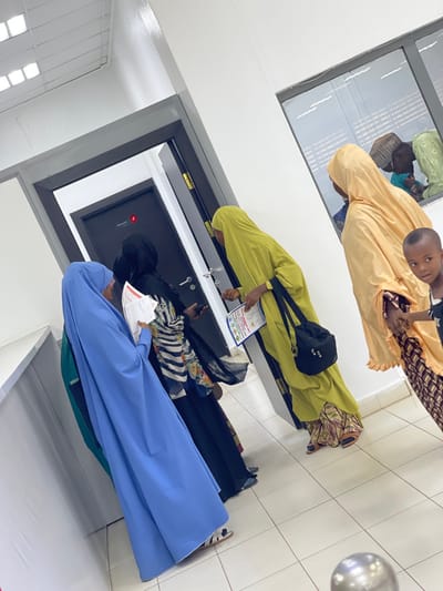 An indoor scene within a building in Zaria, Nigeria, depicts five individuals and reflections of others in what appears to be a waiting or reception area. The environment features white walls, a white tiled floor, and overhead fluorescent lighting. Two dark grey doors with silver handles are visible on the left, one partially open, revealing a darker interior. The visible door displays a small red square emblem and faint text "Opening Hr."
From left to right:
1. A woman, obscured from the waist down, wears a blue abaya and hijab, holding white papers in her hands.
2. Next to her, a woman in a black abaya and hijab, with a striped and floral garment visible underneath, holds a mobile phone in her left hand and papers in her right. She faces the central door.
3. A woman in a lime green abaya and hijab, carrying a black shoulder bag, extends her right hand towards the door frame or a wall-mounted device next to the door. She holds a document with a colorful emblem in her left hand.
4. On the right, a woman in a light orange/peach abaya and hijab stands, holding the hand of a young child.
5. The child, partially visible in the lower right foreground, has short dark hair and wears a dark top, looking directly towards the camera.
A horizontal window on the right wall reflects other individuals, including an adult with a patterned head covering and a child, suggesting additional people are present outside the immediate frame. A metallic spherical object is visible on the floor in the bottom right corner, possibly part of a barrier or fixture.
