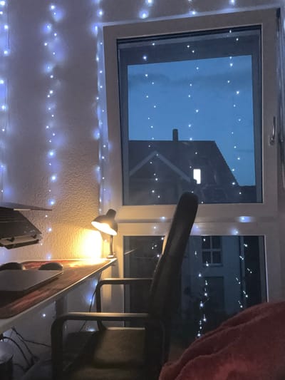 A nighttime interior scene is depicted, likely in Lufingen, Switzerland, with a desk, a chair, a window, and decorative lights. The desk, made of light-colored wood with a dark, patterned mat, holds a laptop and a mouse. To the left of the desk, a black desk lamp with a metallic finish is illuminated, casting a warm yellow glow. The wall behind the desk is textured and adorned with strings of small, blue-white fairy lights.
A dark-colored office chair with a high back is positioned in front of the desk. The scene is dominated by a large window that looks out onto a twilight sky. The sky is a deep blue, dotted with numerous small white lights, some of which appear to be part of the same fairy light string as those indoors, while others might be distant lights or stars. Reflected in the window are silhouettes of a building with a pitched roof and a chimney, and another building with multiple windows.
The overall ambiance is cozy and intimate, suggested by the soft lighting and the decorative string lights. The presence of a laptop and desk lamp implies a workspace, possibly a home office or study area. The view from the window, with the hints of buildings and the twilight sky, grounds the scene in a specific time and place, though the precise location beyond Lufingen, Switzerland is not further specified by the visual elements.