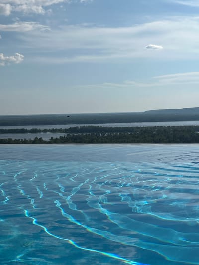 A high-angle, full shot shows an infinity pool in the foreground, its blue water reflecting sunlight and creating rippling patterns. In the middle ground, a wide river is visible, bordered by dense green trees. Beyond the river, a hazy landscape of rolling hills stretches to the horizon under a pale blue sky with scattered white clouds. A dark object, possibly a helicopter or drone, is in the sky above the river. The image appears to be taken from a vantage point overlooking a natural landscape with a body of water, likely during the daytime.