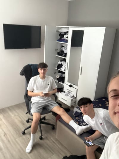 The media file shows two young males in a bedroom. One male is partially visible at the top, his face close to the camera, with a faint smile. He is wearing a white t-shirt. The other male is seated on a black office chair, looking towards the camera with a neutral expression. He is wearing a white t-shirt and grey shorts, and white socks. His left leg is extended. A cell phone is visible in the other male's hand, held up.

In the background, a large white wardrobe with several open compartments is visible. The compartments contain clothing and other items. To the left of the wardrobe, a black flat-screen television is mounted on the wall. The room has light-colored walls and wooden flooring. The scene appears to be indoors, likely in a residential setting. The lighting suggests it could be daytime. No specific weather cues are present. The overall mood appears casual and relaxed. No visible text is present in the image. The location context provided is Belashtitsa, Bulgaria.