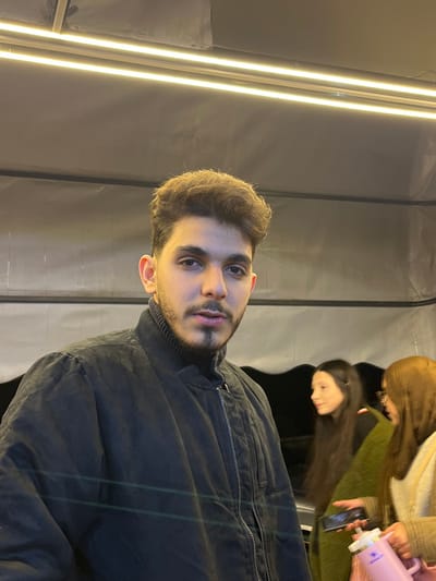 The image depicts a young male in the foreground, centered, looking directly at the camera. He has brown, styled hair and a neatly trimmed mustache and goatee. He is wearing a dark, possibly black or navy blue, bomber-style jacket with a ribbed collar and a visible metallic zipper. Above him, two bright, horizontal LED strip lights are affixed to a dark, possibly fabric or tarp-like, ceiling or canopy.
To the right of the male subject, in the mid-ground and slightly out of focus, are two females. The first female, positioned behind and to the right of the male, has long dark hair and is seen in profile, looking towards the second female. The second female, on the far right, has light brown hair and appears to be holding a dark-colored smartphone in her right hand and a pink thermos-style bottle in her left hand. She is looking downwards towards the items she holds. The background behind the individuals consists of dark material with horizontal creases or structural elements. The scene is located in Al-Muhajerin Municipality, Syria.