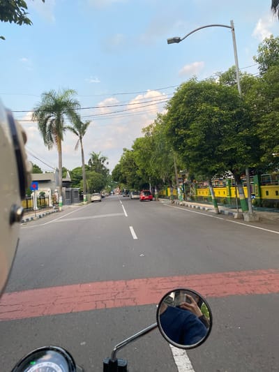 The image presents a first-person perspective from a motorcycle rider traveling along a paved road in Madiun, Indonesia. The scene depicts a clear, bright day, likely late afternoon, with a blue sky dotted with scattered white clouds.

The asphalt road features distinct markings, including a red stripe parallel to a white line on the right side, and a solid white line indicating traffic separation. The road is flanked by abundant greenery, consisting of lush trees and several tall palm trees, suggesting a tropical or subtropical environment. On the right side of the road, beyond the trees, colorful buildings or barriers painted in vibrant yellow and green are visible. Further down the street, other vehicles, including a red car, can be seen. A street lamp with a distinctive curved arm extends over the left side of the road.

In the immediate foreground, the top edge of a helmet or part of the motorcycle's frame is visible at the bottom of the image. A circular rearview mirror on the right prominently reflects the rider's hand and arm, dressed in blue, confirming the vantage point. A portion of the motorcycle's instrument cluster or speedometer is partially visible just below the mirror. Utility poles and power lines are visible against the sky on the left. Further down the road on the left, a blue sign with a white cross, typically denoting a medical facility or first aid, is discernible. The scene conveys a sense of a pleasant journey through a lively urban or suburban setting.