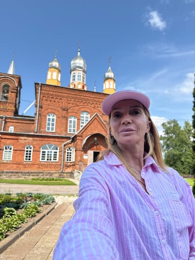 A woman wearing a pink striped shirt and a pink baseball cap takes a selfie in front of a red brick church with silver domes. The church has arched windows and appears to be an Orthodox Christian church. There are green trees and a blue sky in the background. The woman is smiling and looking at the camera. The church is identified as being located in Chudovo, Russia.